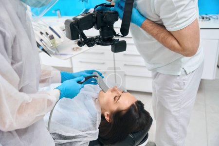 Téléchargez les photos : Médecin avec l'aide d'un assistant photographie les dents des patients, la femme est située dans la chaise dentaire - en image libre de droit