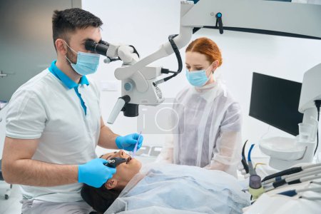 Odontólogo joven en la clínica llena un diente por cliente, un asistente está cerca, el médico utiliza un microscopio en su trabajo