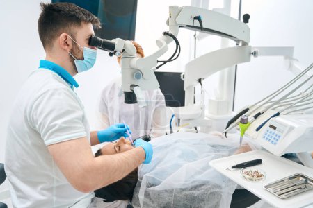 Téléchargez les photos : Dentiste dans une clinique dentaire remplit une dent pour un client, un assistant est à proximité, le médecin utilise un microscope au travail - en image libre de droit
