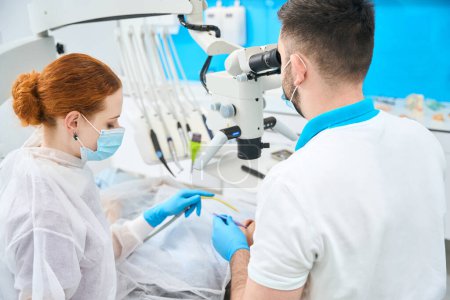 Foto de Médico dentista y su asistente de pelo rojo en su lugar de trabajo, el médico utiliza un microscopio en el trabajo - Imagen libre de derechos