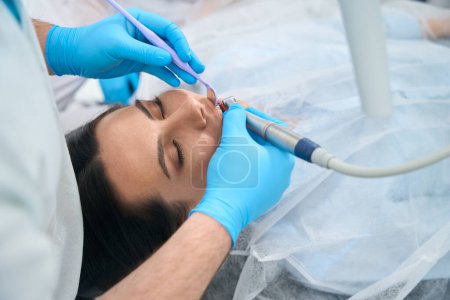 Téléchargez les photos : Médecin dentiste utilise des outils spéciaux et des gants de protection pour le travail, le patient se trouve dans la chaise dentaire - en image libre de droit