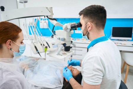 Téléchargez les photos : Homme traite une dent de patients sous un microscope, à côté d'un assistant aux cheveux roux dans un masque de protection - en image libre de droit