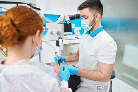 Téléchargez les photos : Médecin traite une jeune dent féminine au microscope, à côté d'un assistant aux cheveux roux dans un masque de protection - en image libre de droit