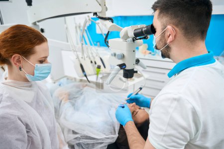 Téléchargez les photos : Médecin dentiste traite une jeune dent féminine au microscope, à côté d'un assistant aux cheveux roux dans un masque de protection - en image libre de droit