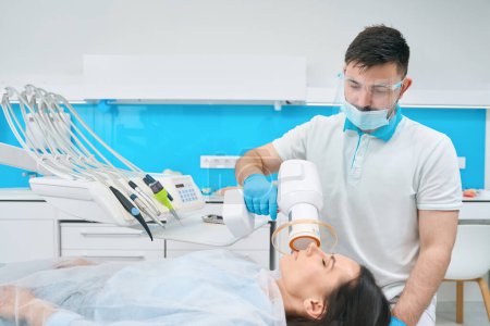 Foto de Médico en el consultorio dental pone un relleno de fotopolímero en una morena, el dentista utiliza una lámpara especial - Imagen libre de derechos