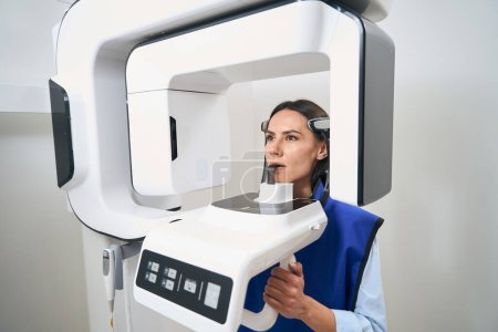 Une patiente brune dans un tablier de protection subit une procédure de numérisation 3D à l'aide d'un équipement moderne