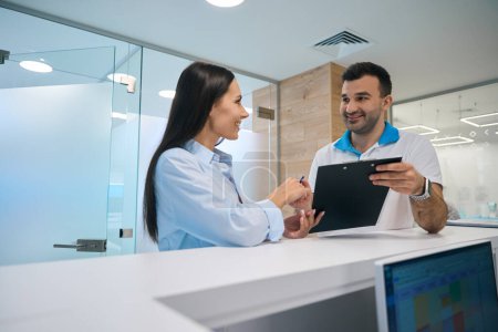 Photo for Smiling man and charming woman are talking at the reception desk in the clinic, the room is clean and bright - Royalty Free Image