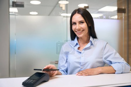 Photo for Pretty woman pays for dental services by phone through the terminal at reception desk, the lady has a snow-white smile - Royalty Free Image