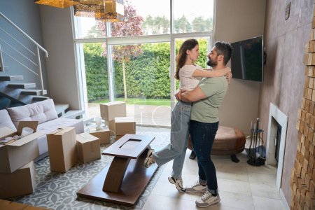 Photo for Handsome man in casual clothes lifting his girlfriend and hugging her in modern room - Royalty Free Image