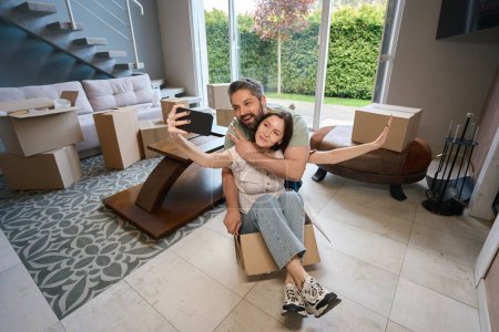 Photo for Pretty female in casual clothes sitting on box and taking selfie with her happy partner indoors - Royalty Free Image