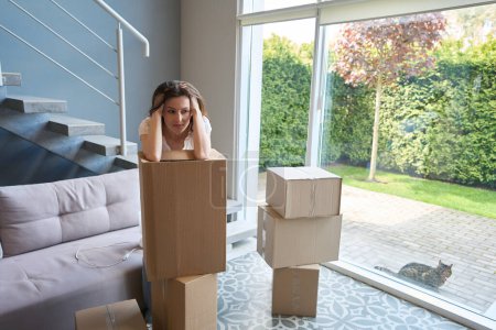 Foto de Hermosa mujer poniendo sus codos en cajas de cartón y pensando mientras está sentada en una habitación moderna - Imagen libre de derechos