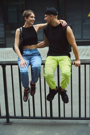 Photo for Cute woman and man in casual clothes sitting on railing and showing affection to each other outside - Royalty Free Image