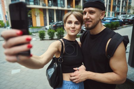 Photo for Beautiful couple in casual clothes making selfie by using cell phone and standing in modern street - Royalty Free Image