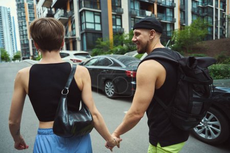 Téléchargez les photos : Homme souriant avec sac à dos regardant sa petite amie pendant qu'ils marchent dans la rue moderne - en image libre de droit