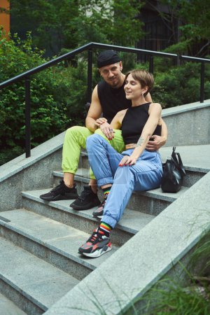 Téléchargez les photos : Jeune homme regardant la caméra tout en étant assis sur les escaliers près de sa petite amie heureuse dans le parc - en image libre de droit