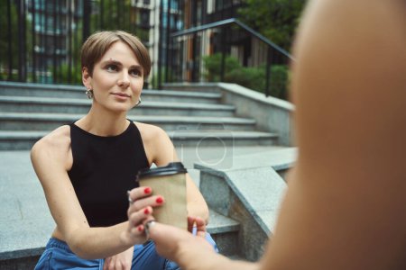 Téléchargez les photos : Femme hipster en vêtements fantaisie recevant tasse de café tout en étant assis sur les escaliers dans la zone urbaine - en image libre de droit