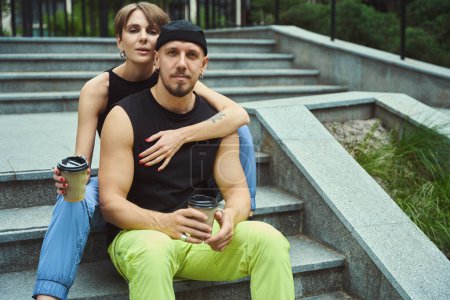 Téléchargez les photos : Beau couple en vêtements décontractés avec tasse de café en regardant la caméra et assis sur les escaliers dans le parc - en image libre de droit