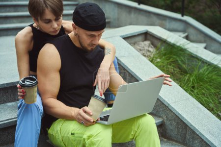 Téléchargez les photos : Jeune femme montrant de l'affection pour son petit ami qui utilise un ordinateur portable tout en étant assis sur les escaliers dans le parc - en image libre de droit