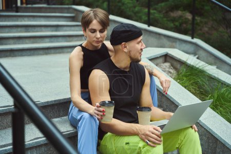 Téléchargez les photos : Bel homme avec ordinateur portable sur ses genoux regardant loin tout en étant assis à sa petite amie à l'extérieur - en image libre de droit