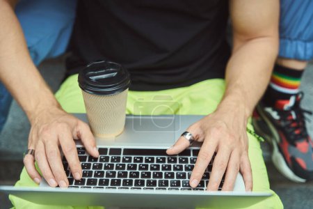 Téléchargez les photos : Vue du dessus sur les mains de l'homme tapant sur ordinateur portable avec une tasse de café dessus tout en étant assis à l'extérieur - en image libre de droit