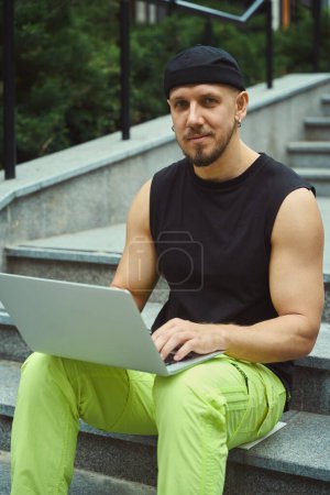 Téléchargez les photos : Homme diligent regardant la caméra tout en utilisant un ordinateur portable et assis sur les escaliers à l'extérieur - en image libre de droit
