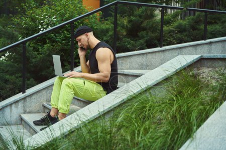 Foto de Freelancer masculino hablando por teléfono celular y trabajando en el portátil mientras está sentado en las escaleras en el parque - Imagen libre de derechos