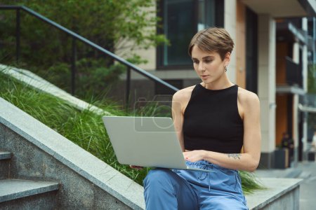 Téléchargez les photos : Jeune femme utilisant ordinateur portable tout en étant assis près de l'herbe dans la rue moderne - en image libre de droit