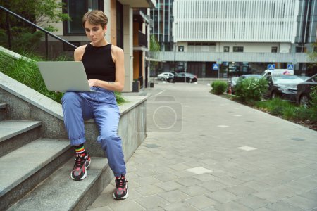 Téléchargez les photos : Jolie femme en vêtements fantaisie à l'aide d'un ordinateur portable tout en étant assis près de l'herbe dans la rue moderne - en image libre de droit