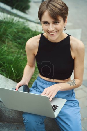Téléchargez les photos : Femme heureuse regardant la caméra tout en utilisant un ordinateur portable et tenant le téléphone portable à l'extérieur - en image libre de droit