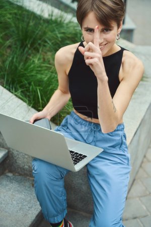 Photo for Attractive female with laptop on her knees looking at camera and pointing up while sitting on modern street - Royalty Free Image