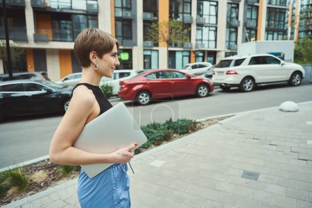 Photo for Young woman in casual clothes holding notebook computer and walking down modern street - Royalty Free Image