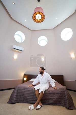 Foto de El huésped del hotel en un albornoz se sienta en una cama grande en un dormitorio acogedor, en el interior hay ventanas redondas - Imagen libre de derechos
