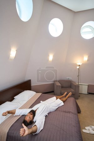 Foto de Barbudo macho se encuentra y se extiende sobre una cama grande en un acogedor dormitorio, la cama está perfectamente hecha - Imagen libre de derechos