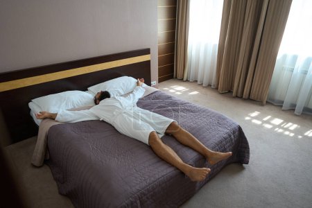 Téléchargez les photos : Homme adulte se trouve sur un grand lit dans une chambre confortable, le lit est soigneusement fait - en image libre de droit