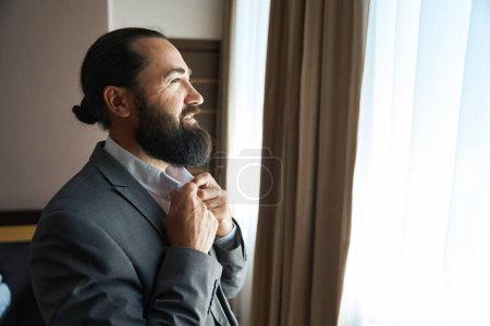 Photo for Handsome man admiring the view from the window of the hotel room, large bed indoors - Royalty Free Image