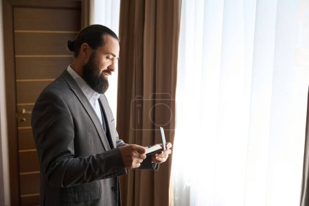 Téléchargez les photos : Homme barbu se tient à la fenêtre et examine un billet, mâle dans une bonne combinaison de voyage - en image libre de droit
