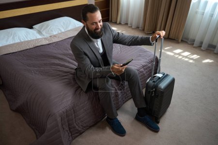 Foto de Barbudo con traje de viaje se sienta en una cama en una habitación de hotel, tiene una pequeña maleta de viaje - Imagen libre de derechos