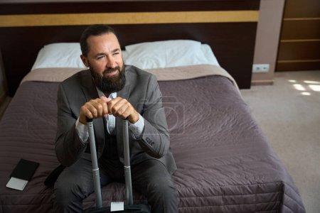 Foto de Barbudo hombre de mediana edad se sienta en una cama en un dormitorio de hotel, tiene una pequeña maleta de viaje - Imagen libre de derechos