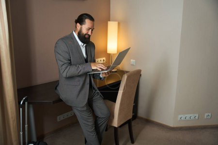Téléchargez les photos : Homme joyeux communique par ordinateur portable dans la zone de travail de la chambre d'hôtel, à côté d'une valise de voyage - en image libre de droit