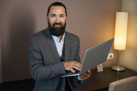 Téléchargez les photos : Voyager mâle communique via ordinateur portable dans la zone de travail de la chambre d'hôtel, un homme en costume de voyage - en image libre de droit