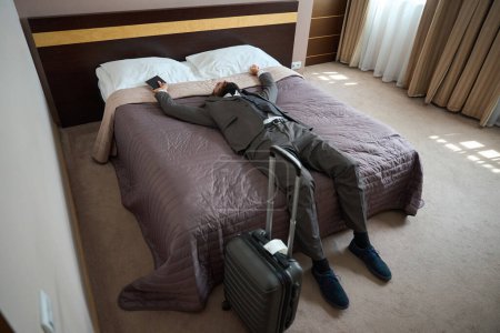 Téléchargez les photos : Voyageur fatigué se repose de la route dans la zone de sommeil de la chambre d'hôtel, homme en costume de voyage solide - en image libre de droit