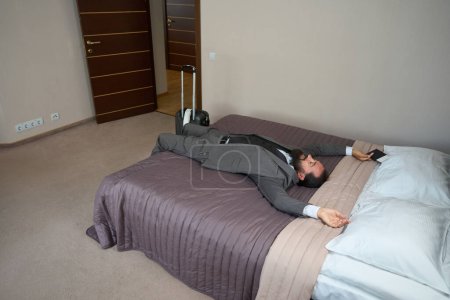 Téléchargez les photos : Homme fatigué se repose de la route sur un grand lit dans une chambre d'hôtel, un homme dans un bon costume de voyage - en image libre de droit