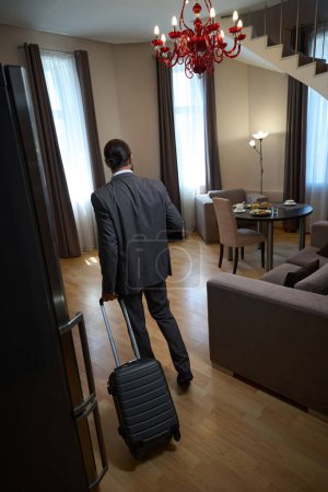 Photo for Man in a business suit checks into a hotel room, he has a small travel suitcase - Royalty Free Image