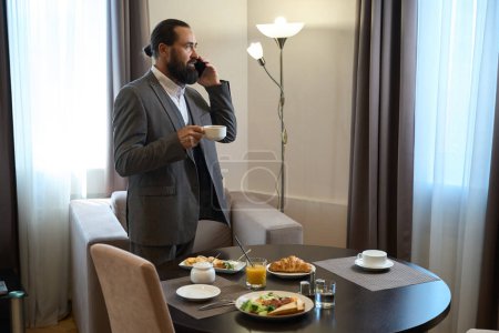 Foto de Empresario hablando por teléfono durante el desayuno, la mesa está puesta para una persona - Imagen libre de derechos