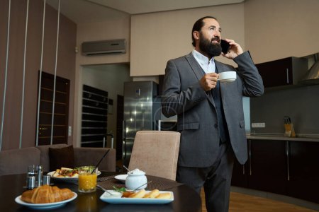 Photo for Male stands at the table with a phone and coffee, the table is set for one person - Royalty Free Image
