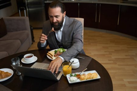 Foto de Hombre barbudo desayuna en una habitación de hotel, un portátil de trabajo está sobre la mesa - Imagen libre de derechos