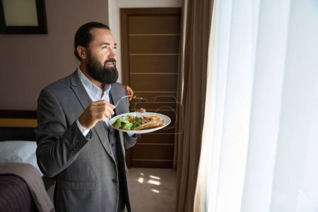 Foto de El hombre barbudo sostiene un plato de comida en sus manos, se para junto a la ventana en una habitación de hotel - Imagen libre de derechos