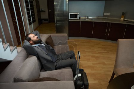 Foto de Un hombre de negocios con un traje de viaje descansa en un sofá suave en una habitación de hotel, tiene una maleta de viaje - Imagen libre de derechos
