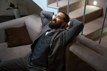 Photo for Satisfied traveler from the road is resting on a soft sofa in a hotel room, wearing a solid travel suit - Royalty Free Image