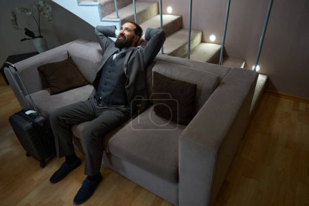 Foto de Hombre satisfecho de la carretera está descansando en un sofá suave en una habitación de hotel, él tiene una maleta de viaje - Imagen libre de derechos